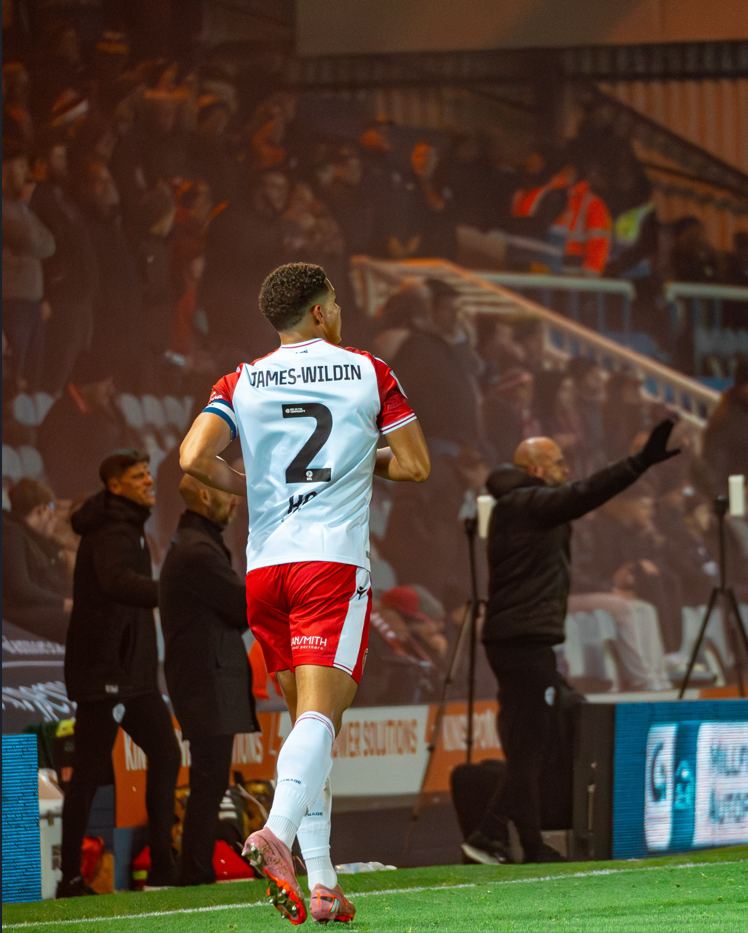 Luther James-Wildin walking off after a goal celebration, stadium crowd behind him